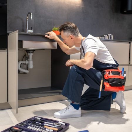 Household repair. Middle-aged man in blue overalls knelt down with hand touching sink carefully examining pipes in kitchen at home